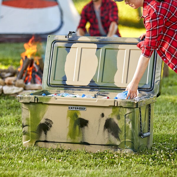 A woman wearing a flannel shirt opening a CaterGator camouflage cooler in the woods.