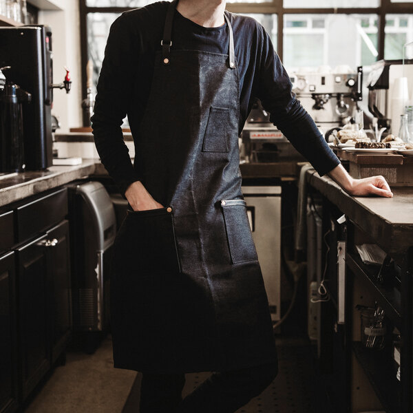 A man in a Hardmill black waxed denim bib apron with 2 pockets standing in a professional kitchen.