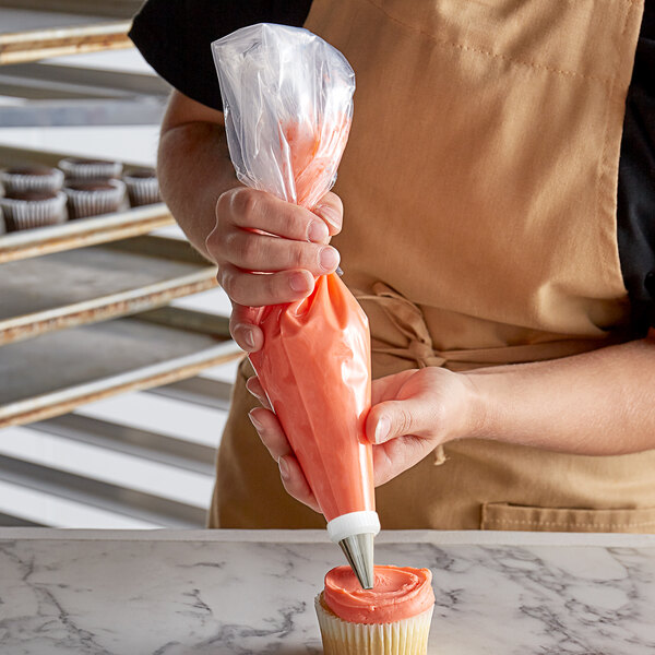 A person in an apron using an Ateco clear disposable pastry bag to pipe pink frosting onto a cupcake.