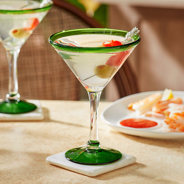 Two Acopa martini glasses with green rims filled with drinks and garnished with olives on a table.