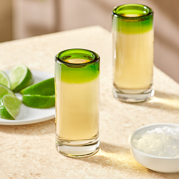 Two Acopa shooter glasses with green rims filled with liquid and lime slices next to a plate of limes.