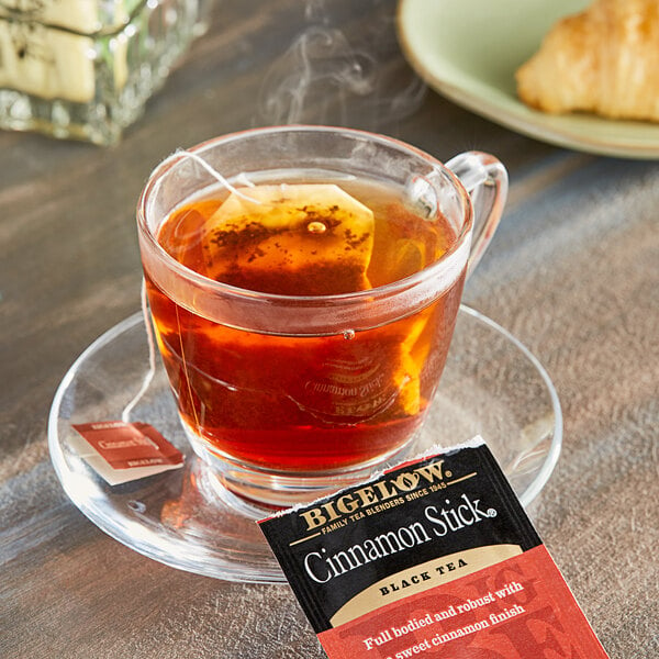 A glass cup of Bigelow Cinnamon Stick tea with a tea bag on a saucer.