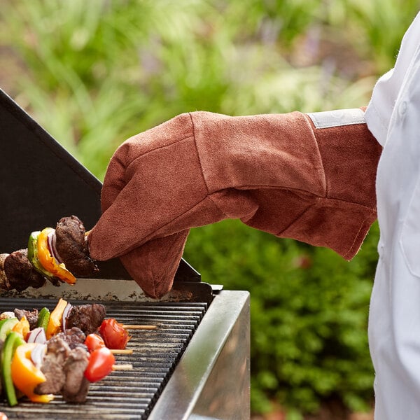 A person wearing an Outset brown leather oven/grill mitt cooking meat on a grill.