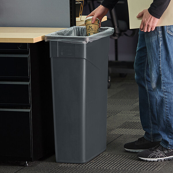 A person standing next to a Carlisle Trimline rectangular trash can holding a coffee cup.