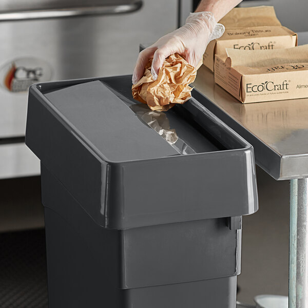 A hand throwing a piece of paper into a Carlisle Trimline trash can with a swing top lid.