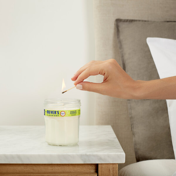 A hand lighting a Mrs. Meyer's Lemon Verbena wax candle in a clear jar.
