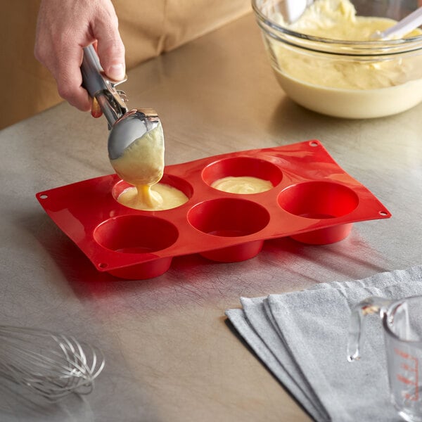 A person using a spoon to pour batter into a Thunder Group red silicone 5 compartment muffin mold on a kitchen counter.