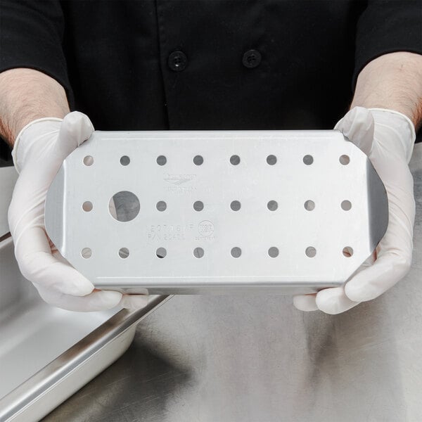A person in gloves holding a Vollrath stainless steel false bottom with holes.
