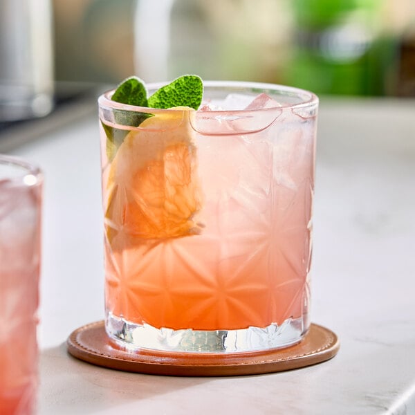 A pink beverage with ice, a lemon slice, and mint leaves served in an 11 oz. patterned rocks glass on a coaster.