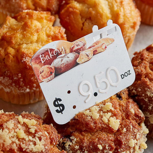 A Ketchum Manufacturing price tag with a number on a card next to muffins in a bakery display.
