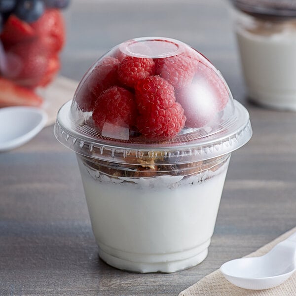 A Choice clear plastic squat cup with yogurt and raspberries with a white spoon on a table.