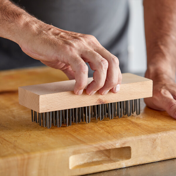 A person's hand using a Carlisle wooden brush with steel bristles to clean a wooden cutting board.