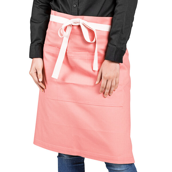 A woman wearing a coral pink Uncommon Chef Moxie waist apron with natural webbing.