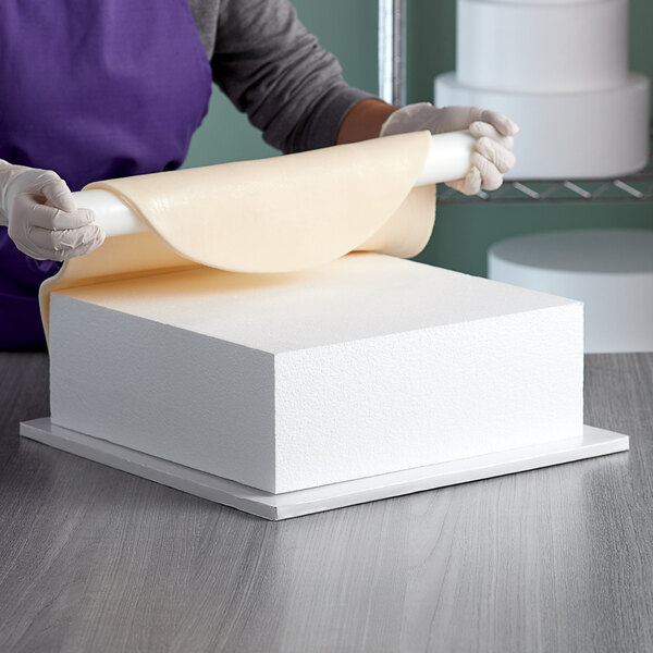 A woman cutting a Baker's Mark foam square cake dummy with a knife.