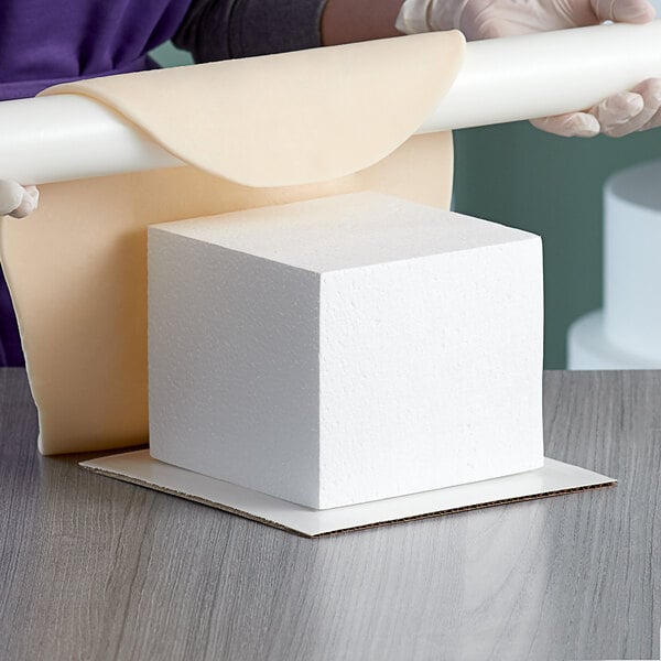A person rolling a white Baker's Mark foam square cake dummy on a table.