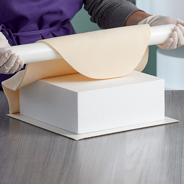 A woman in a purple apron cutting a Baker's Mark foam square cake dummy.