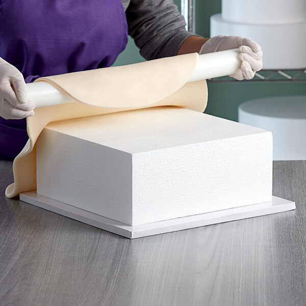 A woman in a purple apron cutting a white square cake dummy.