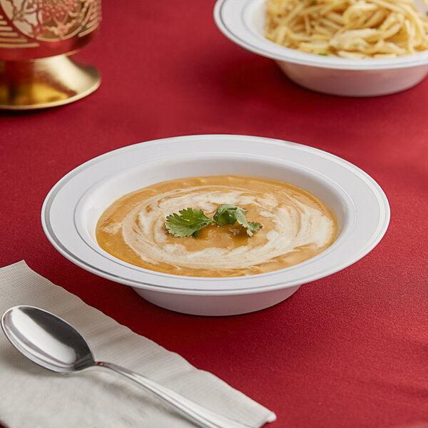 A white plastic bowl of soup with a spoon on a red tablecloth.