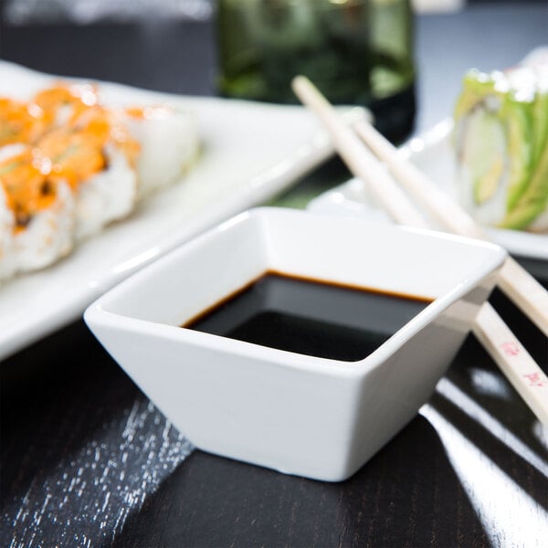 A white square porcelain bowl filled with black liquid next to a plate of sushi with chopsticks.