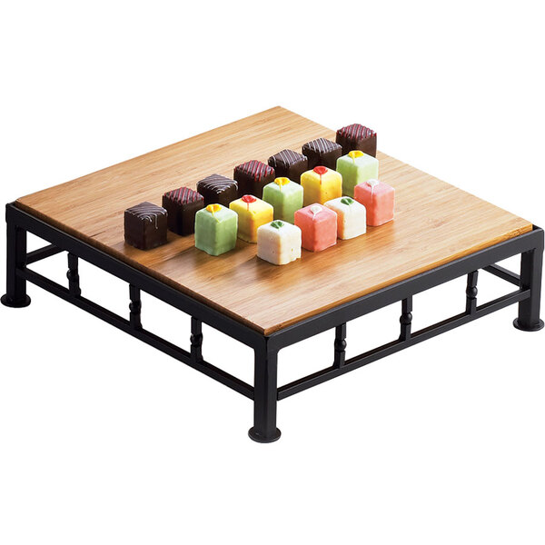 A Cal-Mil black iron square riser with a bamboo top holding a variety of colorful candies on a wood table.