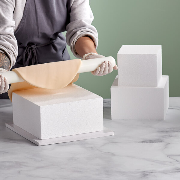 A woman using a rolling pin to shape a Baker's Mark square cake dummy on a white surface.