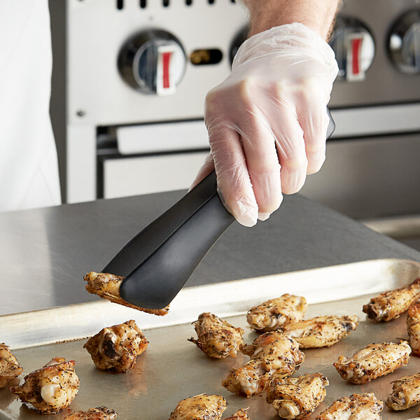 A person wearing plastic gloves using Tablecraft black silicone-coated tongs to pick up a piece of chicken.