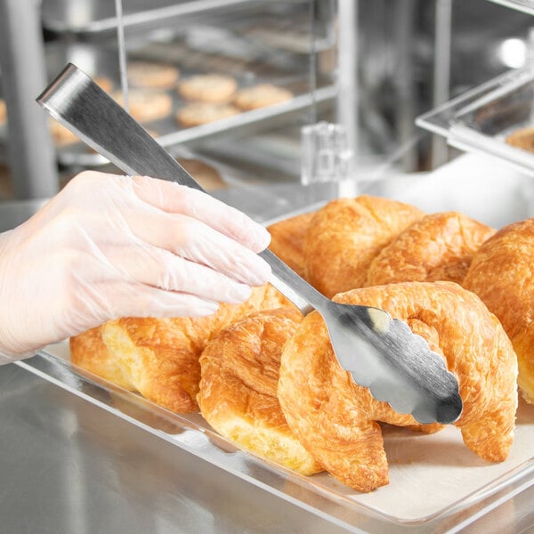 A person in gloves using Carlisle stainless steel scalloped serving tongs to pick up a croissant.