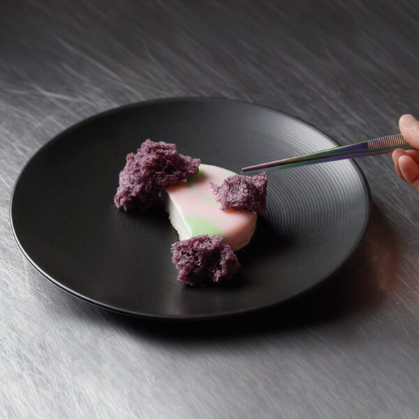 A hand holding a piece of cake with purple crumbs on a Front of the House semi-matte black porcelain plate.