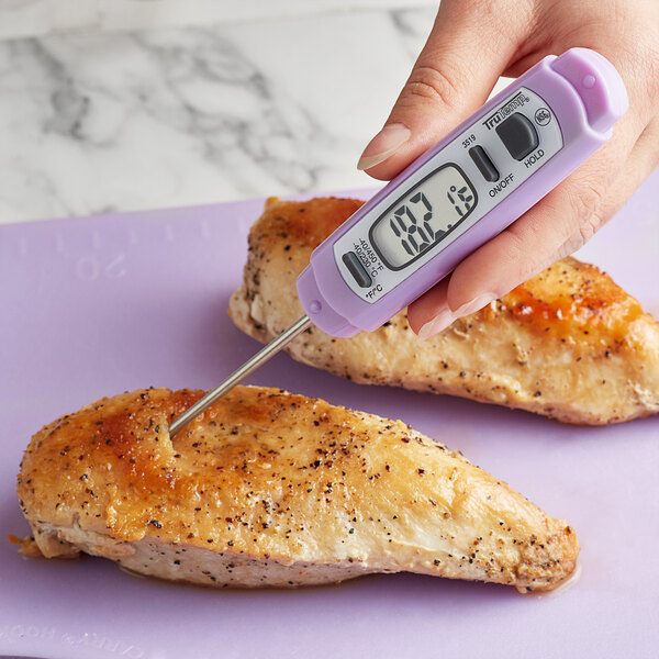 A person's purple hand using a Taylor digital pocket probe thermometer to check the temperature of chicken on a counter.
