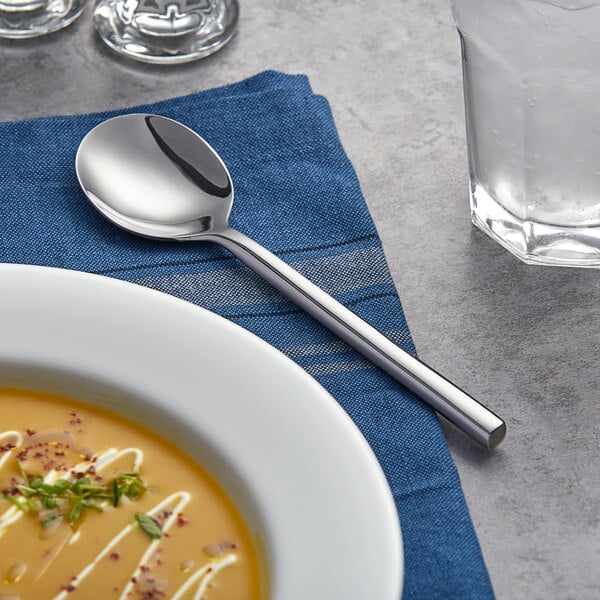 A bowl of soup with a silver Acopa Phoenix bouillon spoon on a plate.