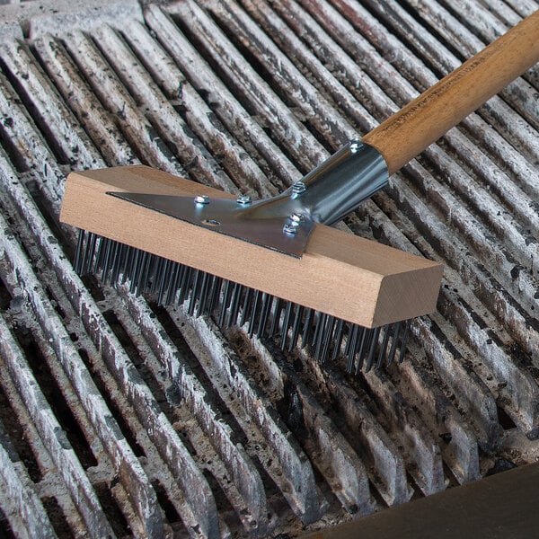 A Carlisle Sparta grill brush with a wooden handle and metal scraper on a grill.