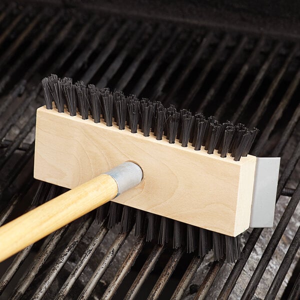 A Carlisle Sparta Broiler Master grill brush with a wooden handle in use.