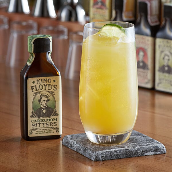 A glass of yellow liquid with a lime slice next to a bottle of King Floyd's Cardamom Bitters.