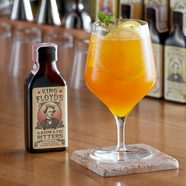 A glass of orange liquid with a slice of lemon and mint in front of a bottle of King Floyd's Aromatic Bitters.