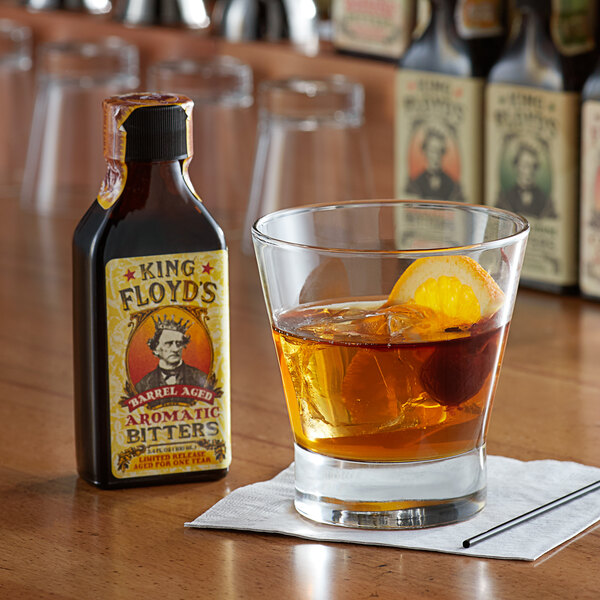 A glass of King Floyd's Barrel Aged Aromatic Bitters next to a glass of brown liquid with an orange slice.