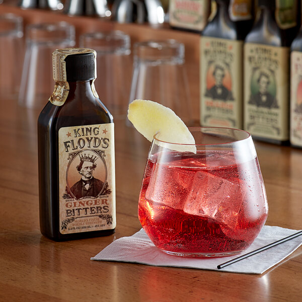 A glass of red liquid with ice cubes and a slice of apple next to a bottle of King Floyd's Ginger Bitters.