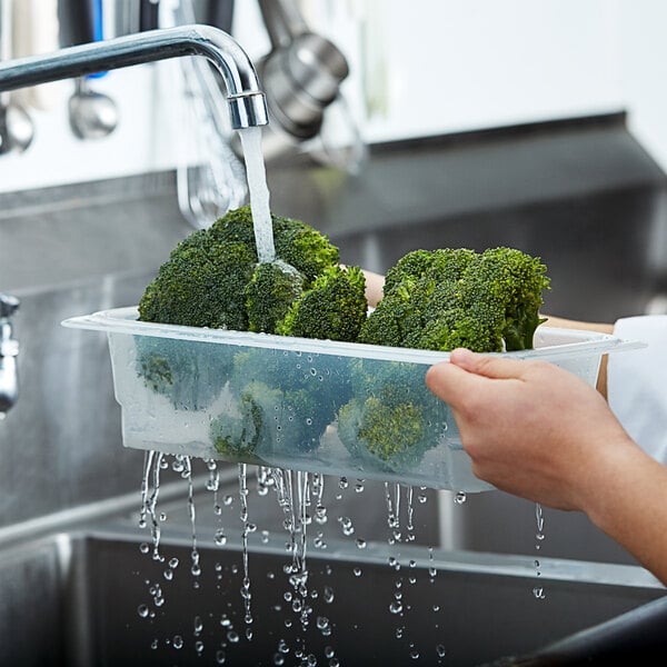 A person washing broccoli in a Cambro plastic food pan colander on a counter.