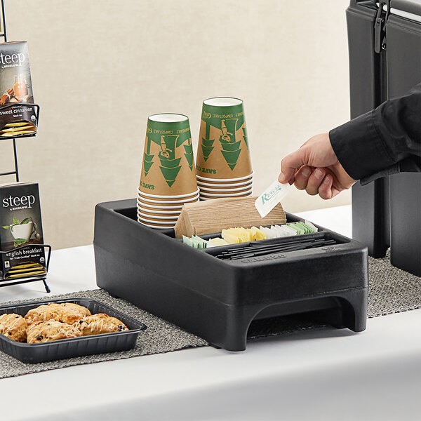 A person holding a black Cambro condiment holder on a table with coffee cups inside.