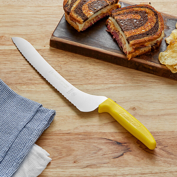 A Dexter-Russell yellow scalloped bread knife next to a sandwich on a cutting board.