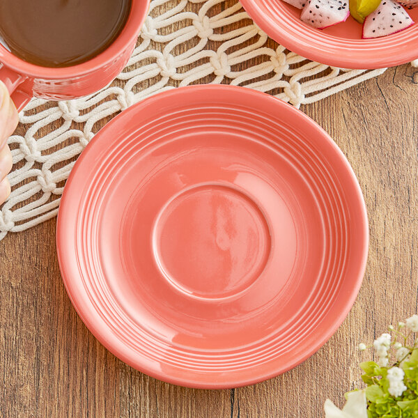 A coral Acopa Capri saucer under a cup with a plant on a wood table.