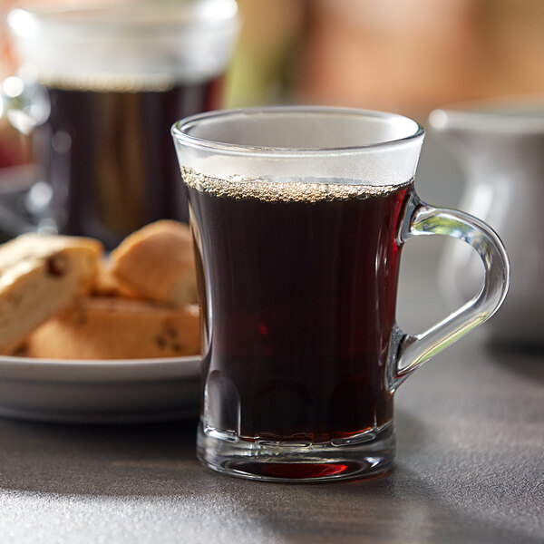 A person holding a Duralex glass espresso mug full of brown coffee with a plate of cookies on the table.