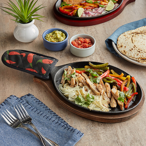 An oval cast iron fajita skillet with food on it next to a bowl of red sauce and a pair of forks.