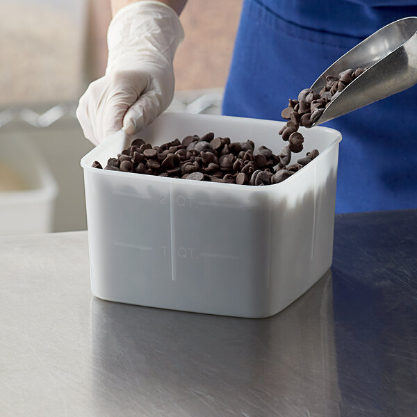 A gloved hand pours chocolate chips from a metal scoop into a white Carlisle food storage container.