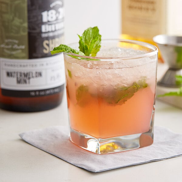 A glass of pink watermelon mint drink with a mint leaf on top on a table with a bottle of 18.21 Watermelon Mint Shrub.