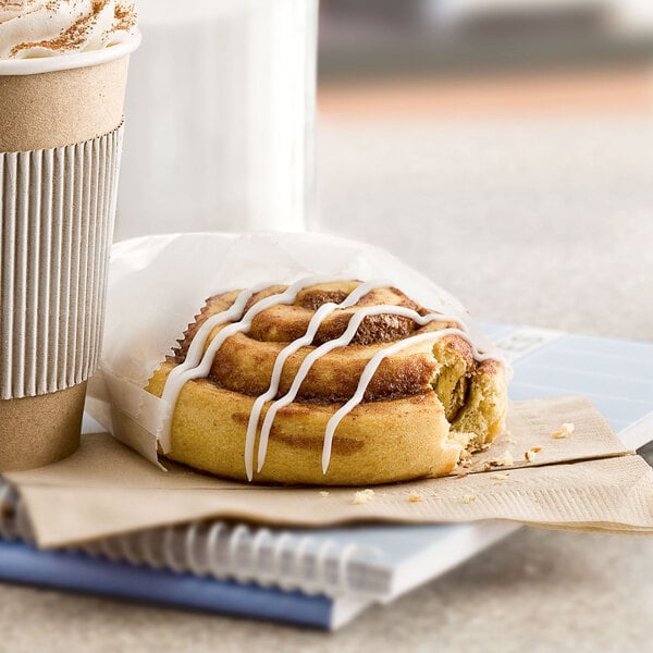 A Rich's cinnamon roll with icing next to a cup of coffee on a table.