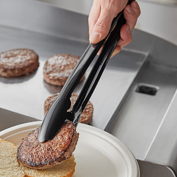 A person using Visions black tongs to place a piece of meat on a burger.