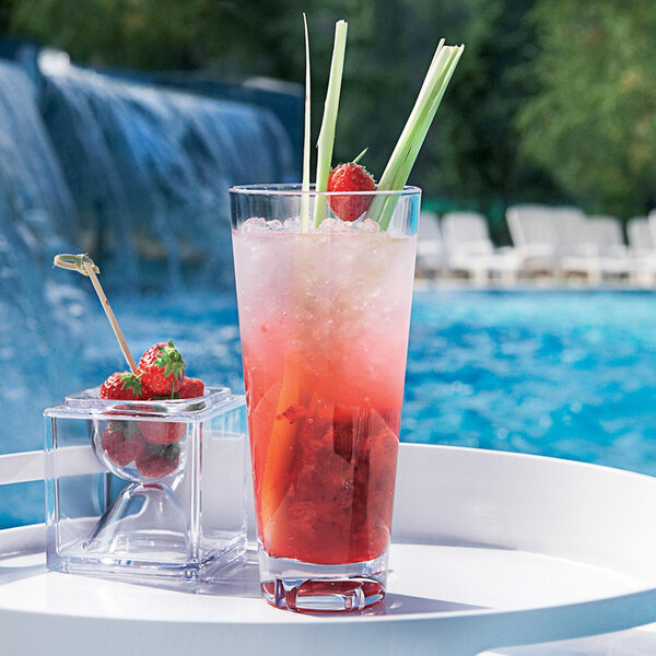 A clear Arcoroc plastic hi ball glass with a red drink and strawberries on a table by a pool.