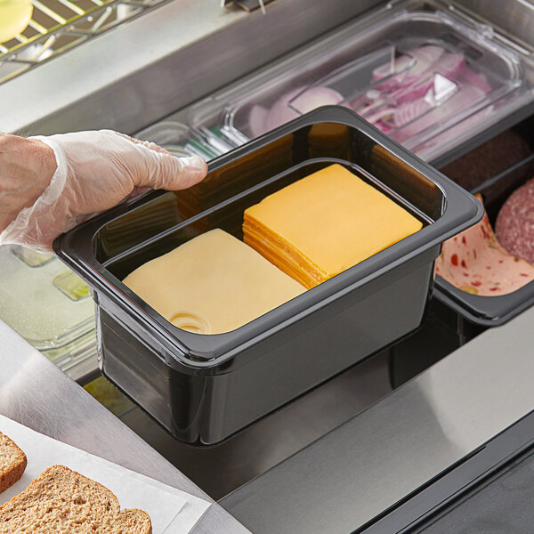 A hand holding a black Vigor polycarbonate food pan filled with cheese on a counter.