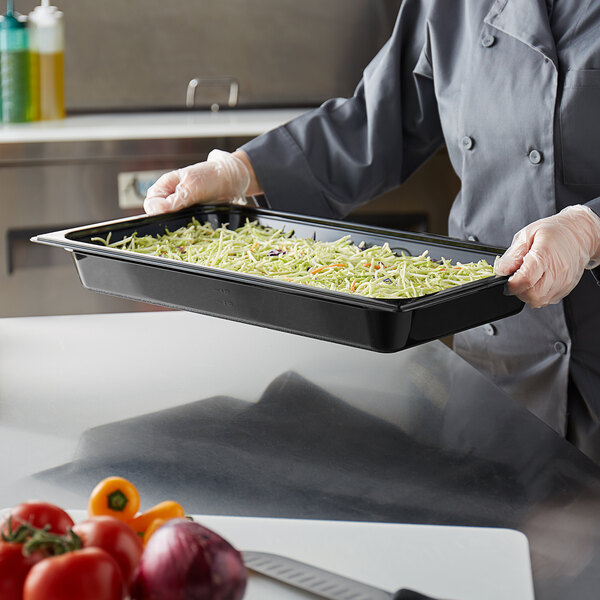 A person in a chef's uniform holding a Vigor black polycarbonate food pan filled with shredded cabbage and broccoli.