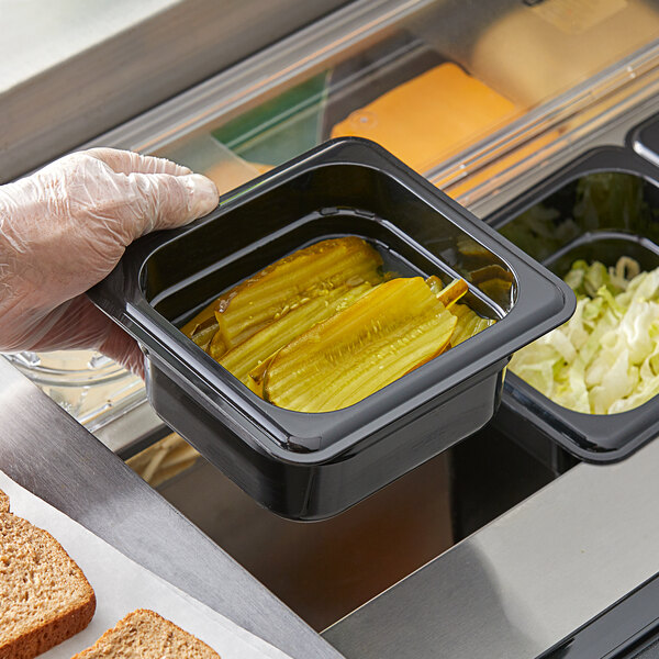 A hand holding a Vigor 1/6 size black plastic food pan of pickles and salad.